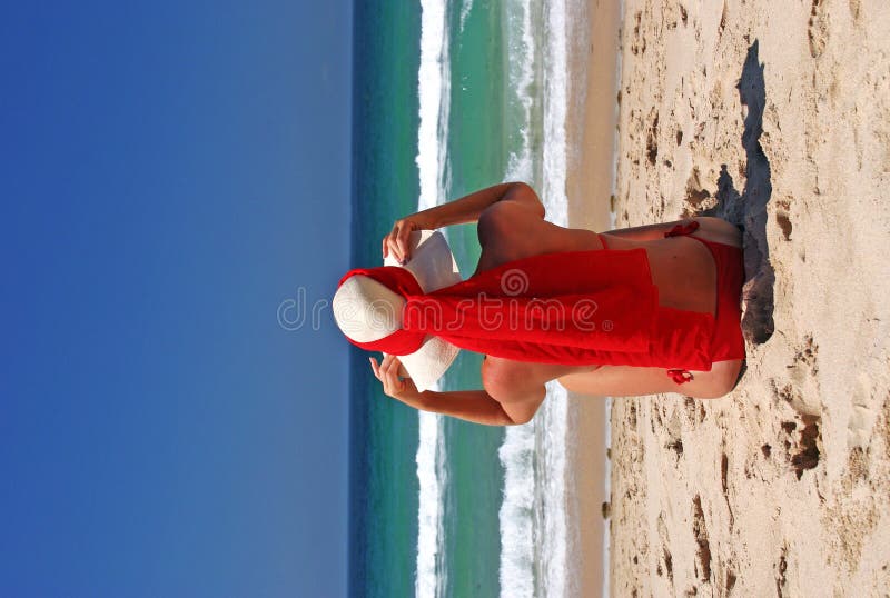 Girl sitting on sandy beach in the sun adjusting hat. Blue sky, blue sea red scarf. Spain.