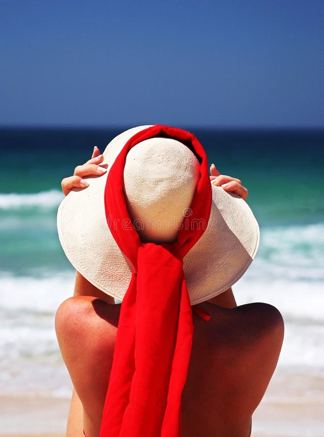 Girl sitting on sandy beach in the sun adjusting hat. Blue sky, blue sea red scarf. Spain.