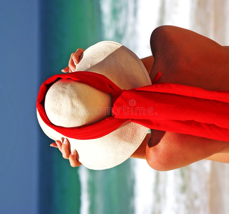 Girl sitting on sandy beach in the sun adjusting hat. Blue sky, blue sea red scarf. Spain.