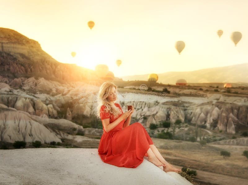 A girl sitting on the top of a cliff with a glass of Turkish tea at dawn with a view of the mountains of Cappadocia and balloons i stock photos A girl sitting on the top of a cliff with a glass of Turkish tea at dawn with a view of the mountains of Cappadocia and balloons i stock photos