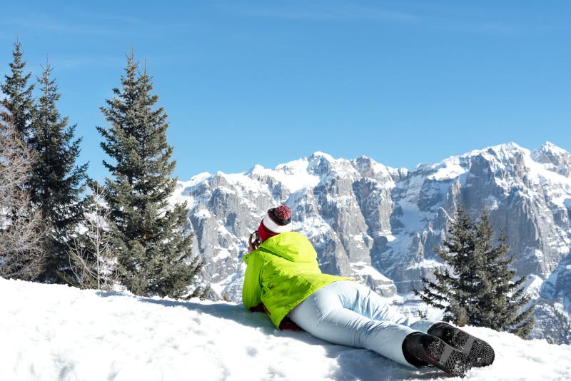 Girl snowboarder in a green jacket lies on a slope and looks at the mountains