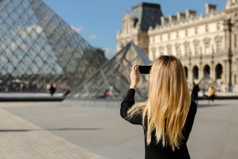 Girl standing near Louvre and glass pyramind in black dress in Paris, talking photo by smartphone.