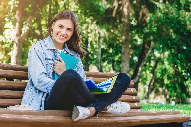 Young girl student sits on a bench in the park and holds books, notebooks and folders. Girl teaches lessons in the park and smiling
