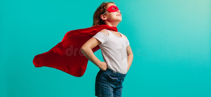 Girl child wearing red superhero costume standing with her hands on hips in studio. Girl in red cape and mask looking away on blue background. Girl child wearing red superhero costume standing with her hands on hips in studio. Girl in red cape and mask looking away on blue background