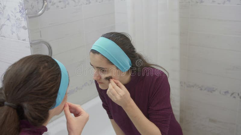 Girl takes off the cosmetic mask from her face in the bathroom and contentedly looks at herself in the mirror