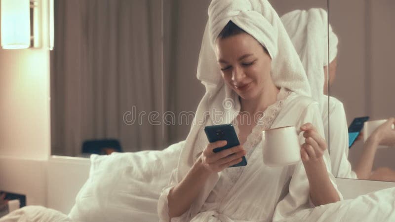 A girl with a towel on her head drinks coffee and chats on a smartphone in the bedroom at night