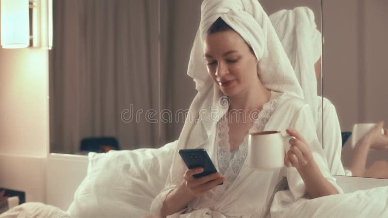 A girl with a towel on her head drinks coffee and chats on a smartphone in the bedroom at night