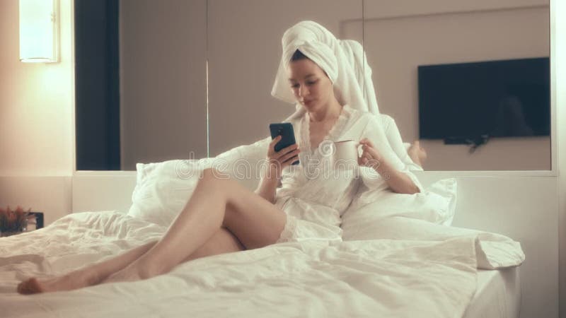 A girl with a towel on her head drinks coffee and chats on a smartphone in the bedroom at night