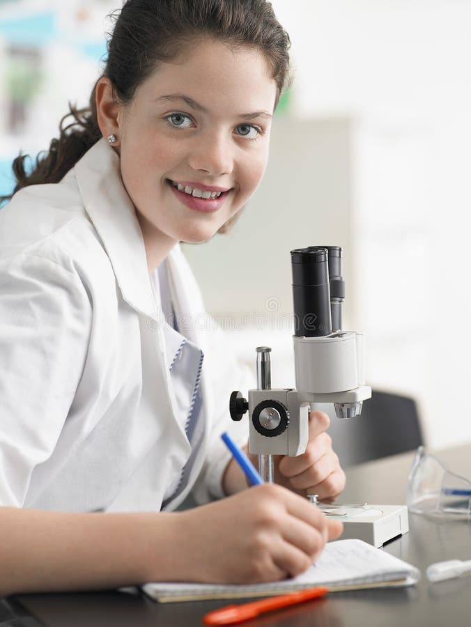 Portrait of happy teenage girl using microscope and taking notes in laboratory