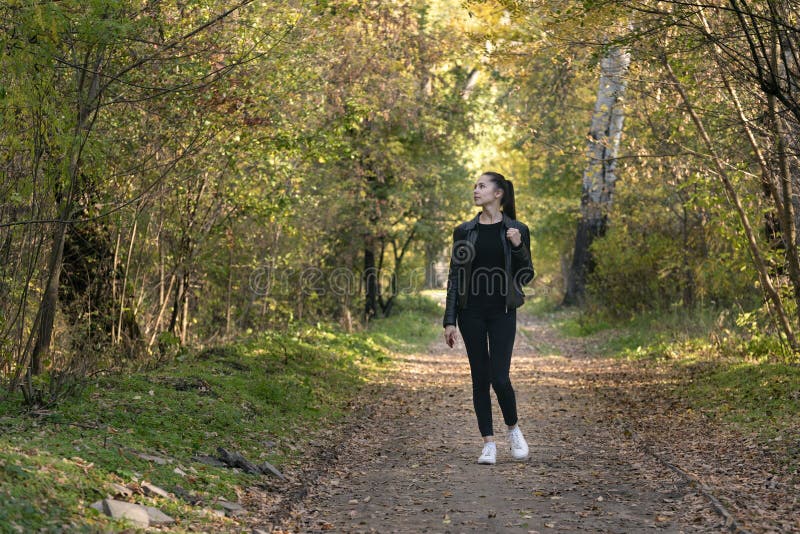Girl walks in the autumn park. Young beautiful woman in black clothes in the forest. Alley, promenade