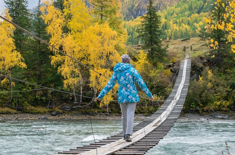 Girl in a warm jacket goes on a wooden suspension bridge on the other side of the turquoise river. Autumn mountain landscape w stock image Girl in a warm jacket goes on a wooden suspension bridge on the other side of the turquoise river. Autumn mountain landscape w stock image
