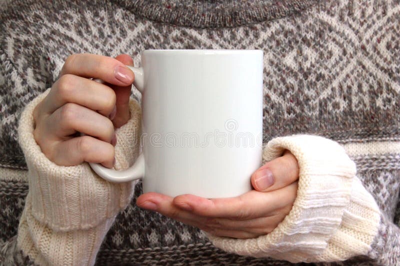 Girl in a warm sweater is holding white mug in hands.