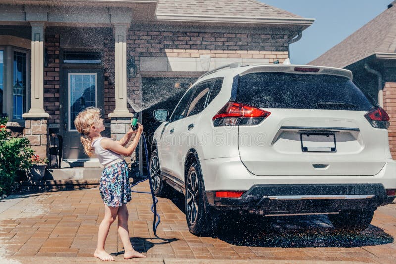 Cute preschool little Caucasian girl washing car on driveway in front house on sunny summer day. Kids home errands duty chores responsibility concept. Child playing with hose spraying water