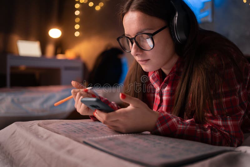 Girl wearing headphones studying using phone while lying on bed. Cute woman student using smartphone tech listening