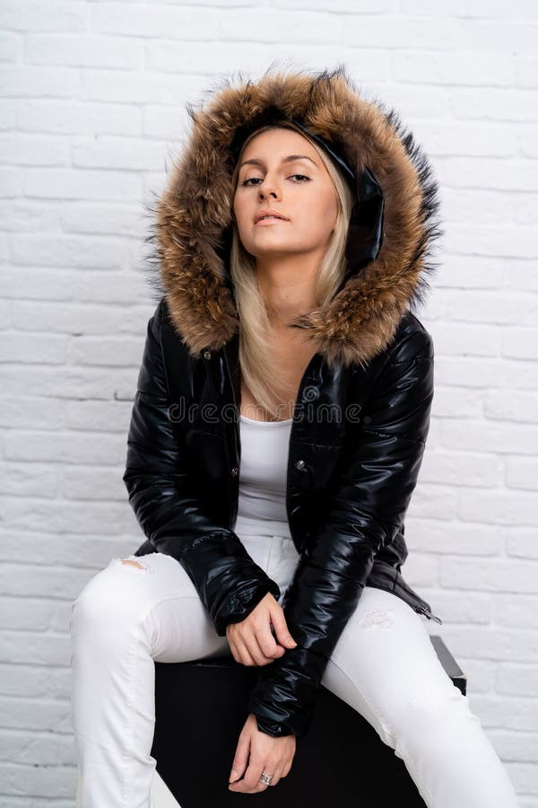 A girl in white jeans, a white blouse and a black shiny jacket with a fur hood on a white background. royalty free stock photography