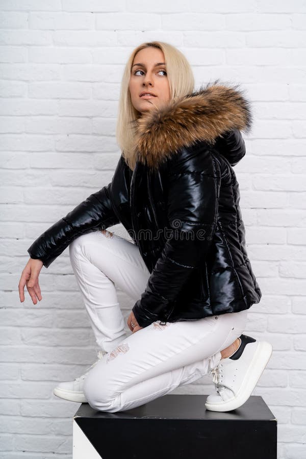 A girl in white jeans, a white blouse and a black shiny jacket with a fur hood on a white background. stock image