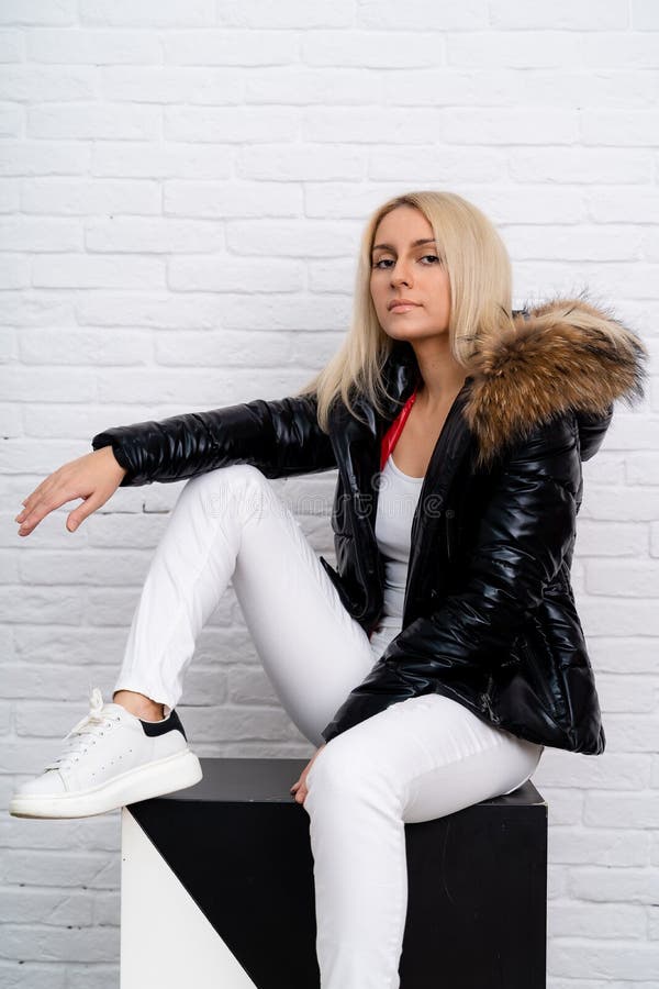 A girl in white jeans, a white blouse and a black shiny jacket with a fur hood on a white background. royalty free stock image
