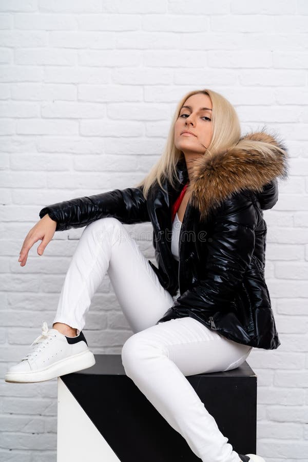 A girl in white jeans, a white blouse and a black shiny jacket with a fur hood on a white background. royalty free stock image