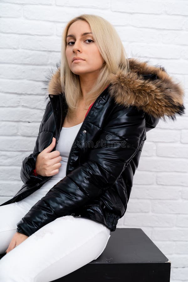 A girl in white jeans, a white blouse and a black shiny jacket with a fur hood on a white background. stock photography