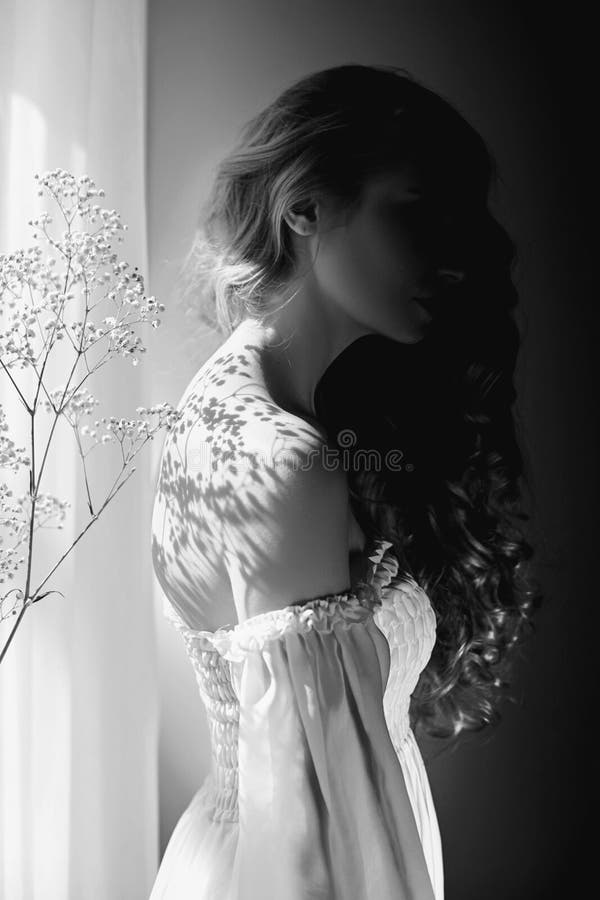 Girl white light dress and curly hair, portrait of woman with flowers at home near window, purity and innocence. Curly blonde