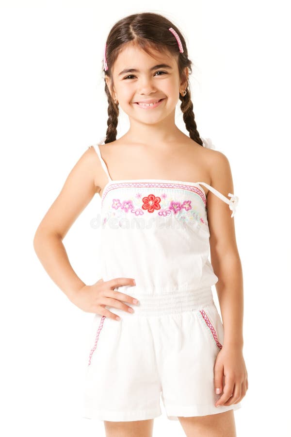 Cute girl in white overall with floral print, pigtails on brunette hair, standing on white background with hand on her belt and smiling