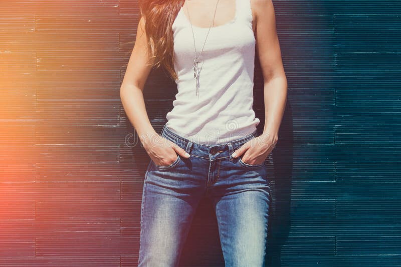 Girl in white tank shirt and blue jeans outdoor summer day middle body lean on tiled wall