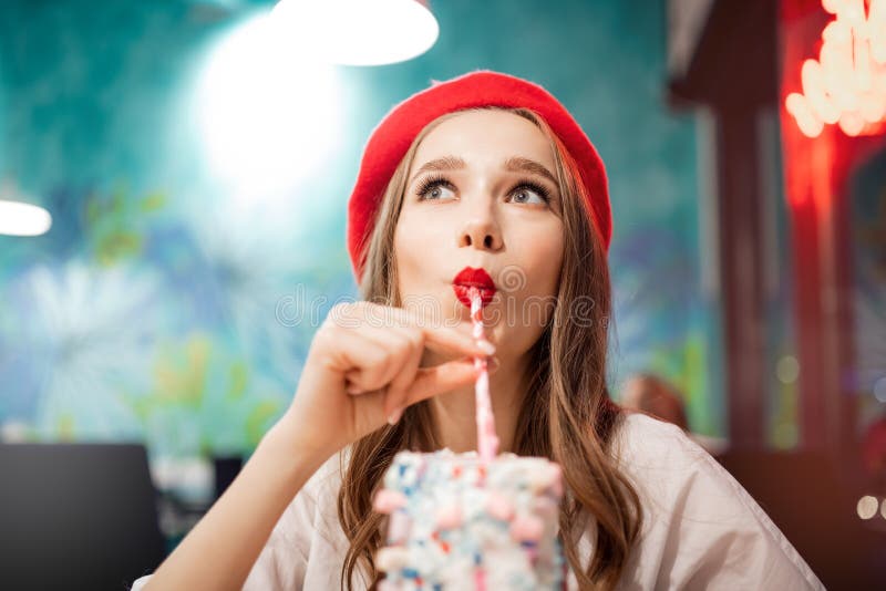 Girl with white teeth, beautiful smile in red beret, France drinks pink strawberry milkshake from straw