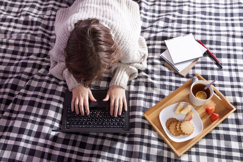 Girl in a white woolen sweater working on a laptop. tray with a cup of tea and a plate with ginger biscuits and an apple on a chec stock photo Girl in a white woolen sweater working on a laptop. tray with a cup of tea and a plate with ginger biscuits and an apple on a chec stock photo