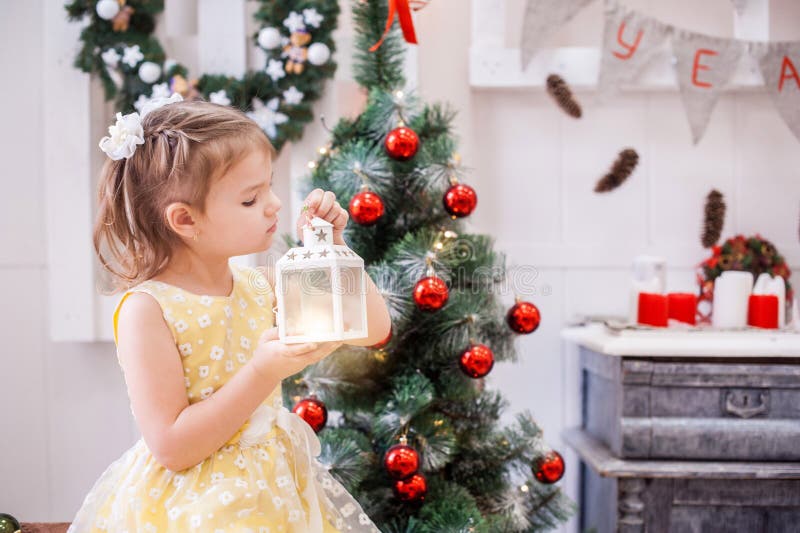 Girl in yellow dress in front of tree holds the Christmas lantern