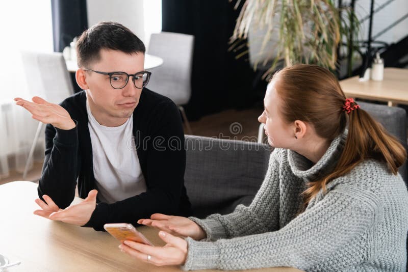 Girlfriend asking for explanation of her boyfriend sitting on a couch in the cafe stock photo Girlfriend asking for explanation of her boyfriend sitting on a couch in the cafe stock photo