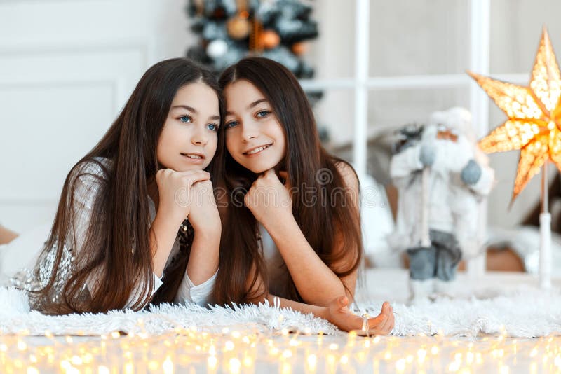 Girls twins in front of the fir-tree. New year`s eve. Christmas. Cozy holiday at the fir-tree with lights stock image Girls twins in front of the fir-tree. New year`s eve. Christmas. Cozy holiday at the fir-tree with lights stock image