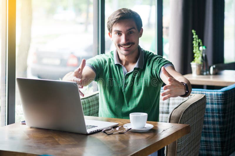 Give me hug. Young happy businessman in green t-shirt sitting looking at camera with toothy smile and want to emrace. business and freelancing concept. indoor shot near big window at daytime