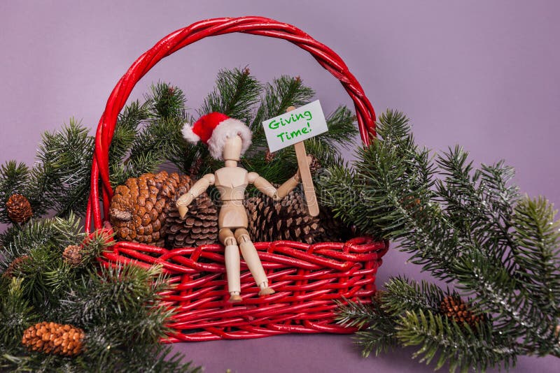 Giving time message held by wooden jointed manikin doll sitting in red basket full of pinecones and pine garland wearing Santa hat royalty free stock image Giving time message held by wooden jointed manikin doll sitting in red basket full of pinecones and pine garland wearing Santa hat royalty free stock image
