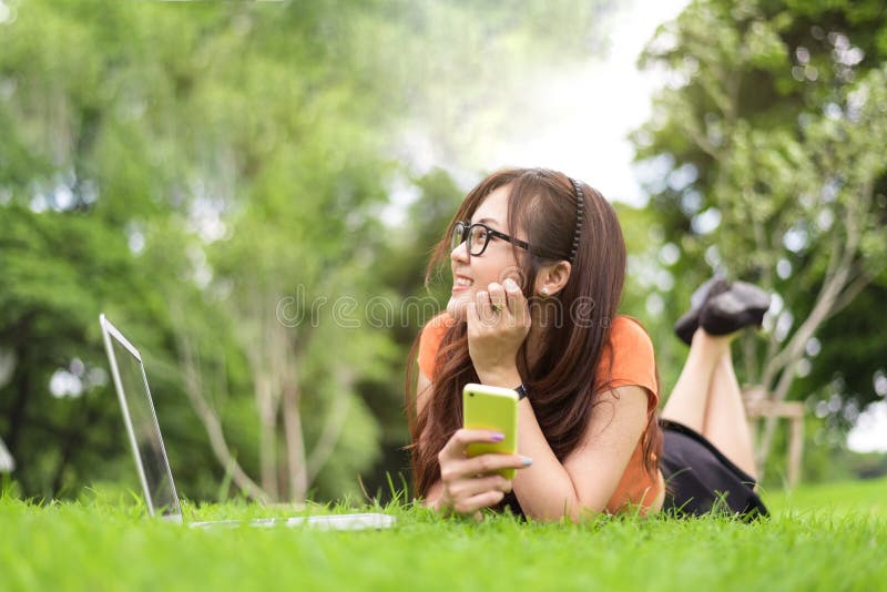 Gl?ckliche Asiatin, die dazu im Park mit Smartphone stillsteht und schaut Leute und Lebensstilkonzept Technologie- und Sch?nheits