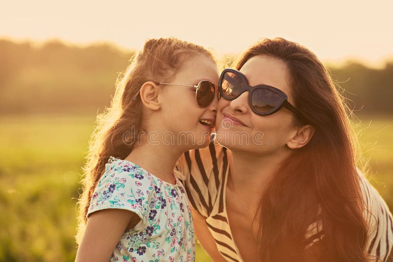 Gl?ckliches Modekinderm?dchen, das ihre Mutter das Geheimnis in der modischen Sonnenbrille in der Profilansicht fl?stert und auf lizenzfreies stockfoto Gl?ckliches Modekinderm?dchen, das ihre Mutter das Geheimnis in der modischen Sonnenbrille in der Profilansicht fl?stert und auf lizenzfreies stockfoto