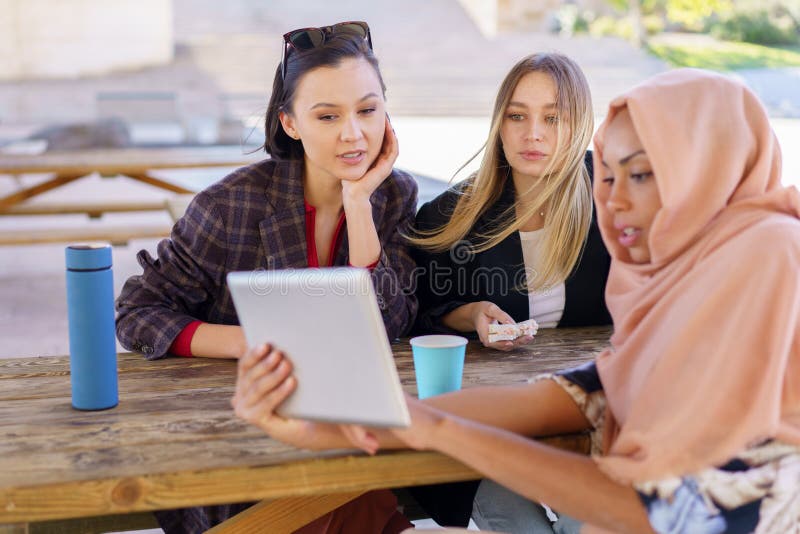 Glad att kvinnor delar på tabletten under kaffeuppehåll i kafé. Grupp av positiva unga multietniska kvinnor som ler och dricker kaffe för att gå medan de tittar på rolig film på surfplattor under inbrott i utomhuscaféet i parken