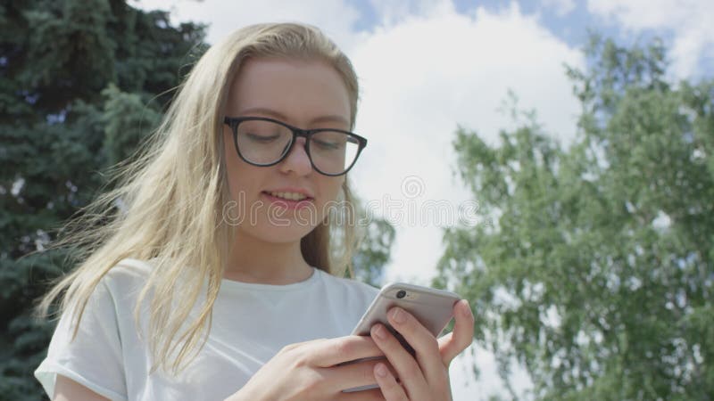 Glad beskådad flicka med blonda hårlekar som håller mobiltelefon i händerna och svarar på meddelandet om solig sommar