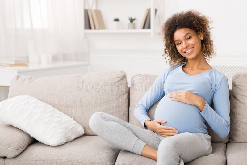 Glad gravid kvinna som sitter på soffan och tar hand om hennes mage
