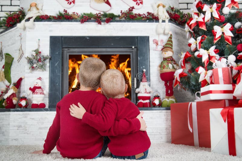 Glad jul och lyckliga ferier! Två bröder som sitter på golvet i vardagsrummet och blicken på branden i spisen I a royaltyfri foto Glad jul och lyckliga ferier! Två bröder som sitter på golvet i vardagsrummet och blicken på branden i spisen I a royaltyfri foto