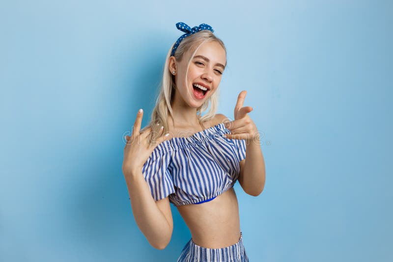 Glad och upphetsad härlig blond flicka i sommardräkt och att peka på kameran som står över blå bakgrund arkivbild Glad och upphetsad härlig blond flicka i sommardräkt och att peka på kameran som står över blå bakgrund arkivbild