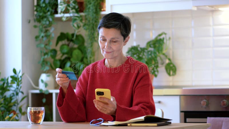 Smiling mature woman makes online payment using smartphone and debit or credit card at home with tea stock video