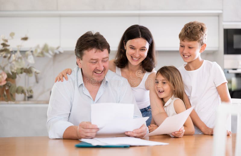 Joyful adult man examining documents while sitting at table in kitchen with his family. Joyful adult man examining documents while sitting at table in kitchen with his family