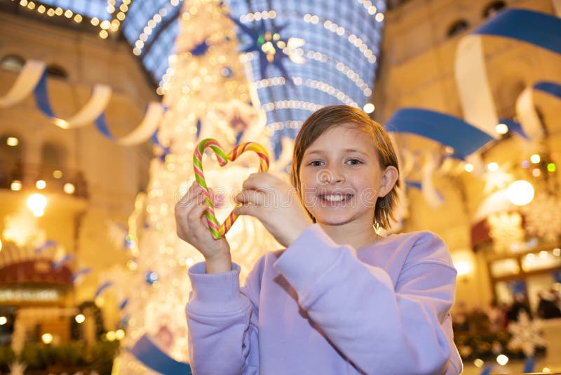 happy child holding heart shaped candy stands in front of christmas decorated tree in winter at mall in evening. happy child holding heart shaped candy stands in front of christmas decorated tree in winter at mall in evening