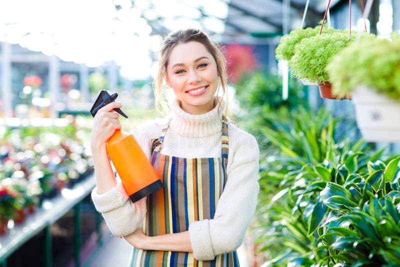 Gladlynt attraktivt kvinnaträdgårdsmästareanseende i orangeri med vattenpulverizeren royaltyfri fotografi Gladlynt attraktivt kvinnaträdgårdsmästareanseende i orangeri med vattenpulverizeren royaltyfri fotografi