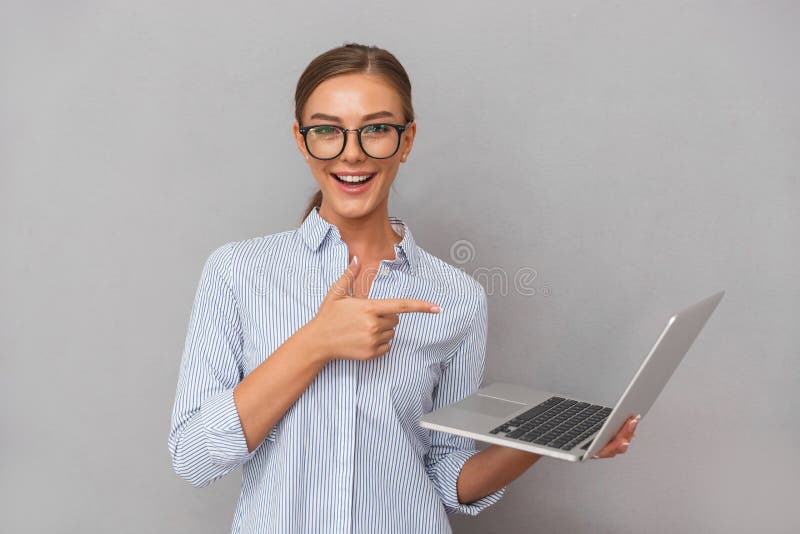 Gladlynt lyckligt posera för ung kvinna för affär som isoleras över grå väggbakgrund genom att använda att peka för bärbar datord