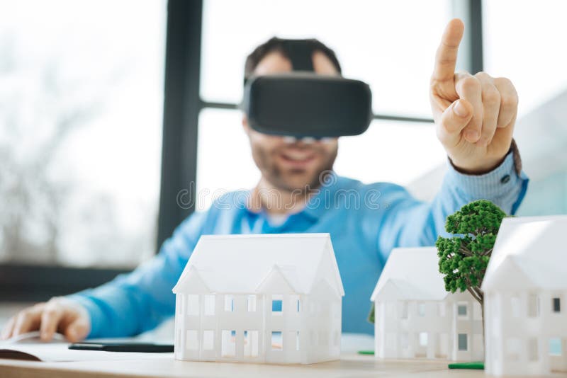 Gladlynt tekniker i VR-hörlurar med mikrofon som pekar på något med fingret. Ny erfarenhet Charmigt sammanträde för upptaktbarntekniker på skrivbordet mycket av den husmodeller och provningen en VR-hörlurar med mikrofon, medan peka på något med hans finger