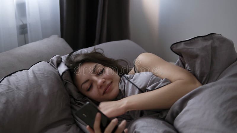 Gladlynt ung brunettflicka som ligger under filten och ser till hennes mobiltelefon i säng Dagtid som kopplar av tid, tar
