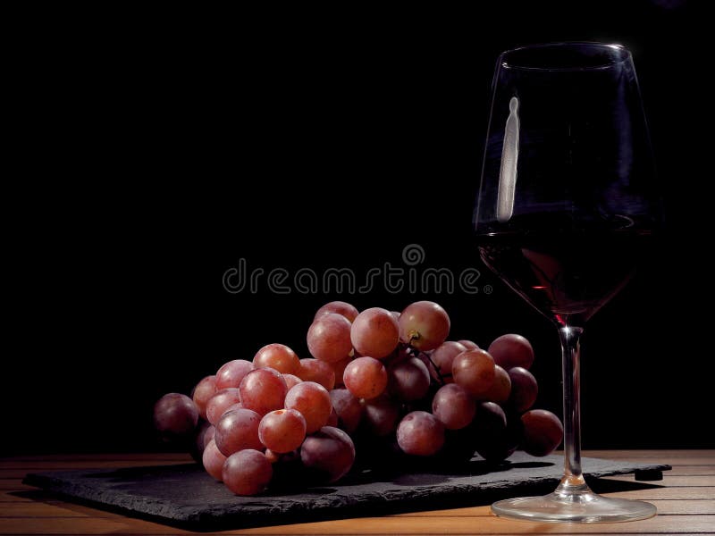 Glas Rotwein und Gruppe von roten frischen organischen Trauben auf einer schwarzen Schieferplatte und -Holztisch stockbild