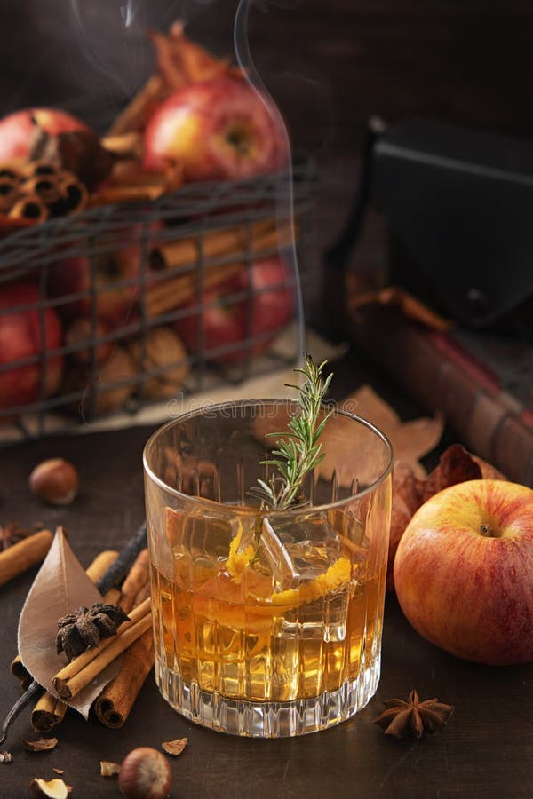 Glass of alcoholic whiskey drink with a branch of rosemary with smoke and apples, cinnamon sticks. Concept of magical autumn