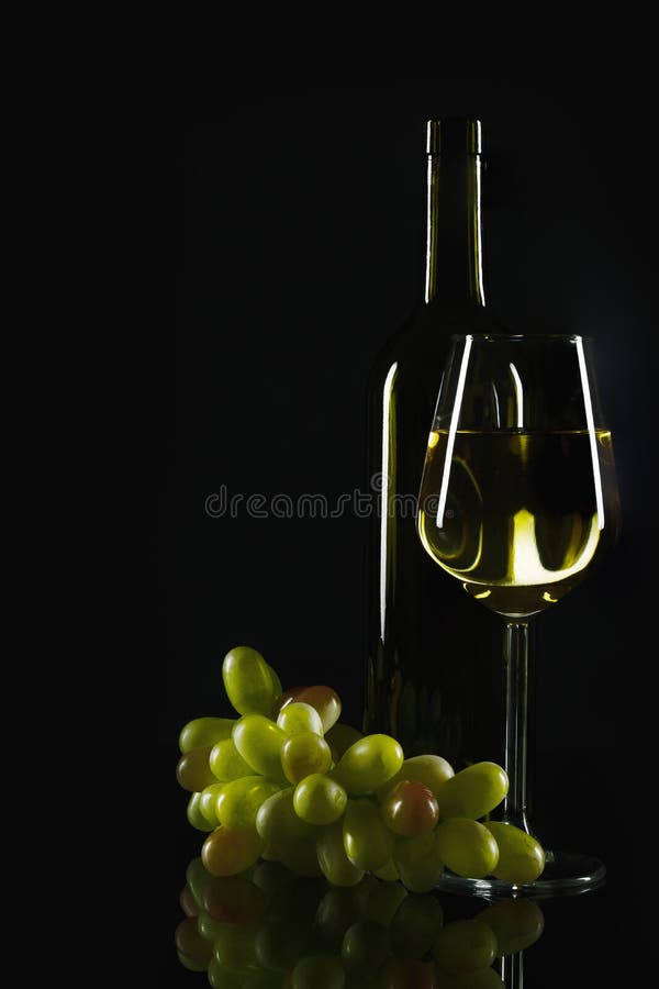 Glass and bottle of white wine with fresh green grapes with reflection on dark background